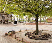 Ein Spielplatz mit einer Holzspielburg, einer Rutsche und einem Baum in der Mitte. Um den Baum liegen Holzstämme. Im Hintergrund sind Schaukeln und eine sandige Fläche. Die Umgebung ist grün und einladend für Kinder.