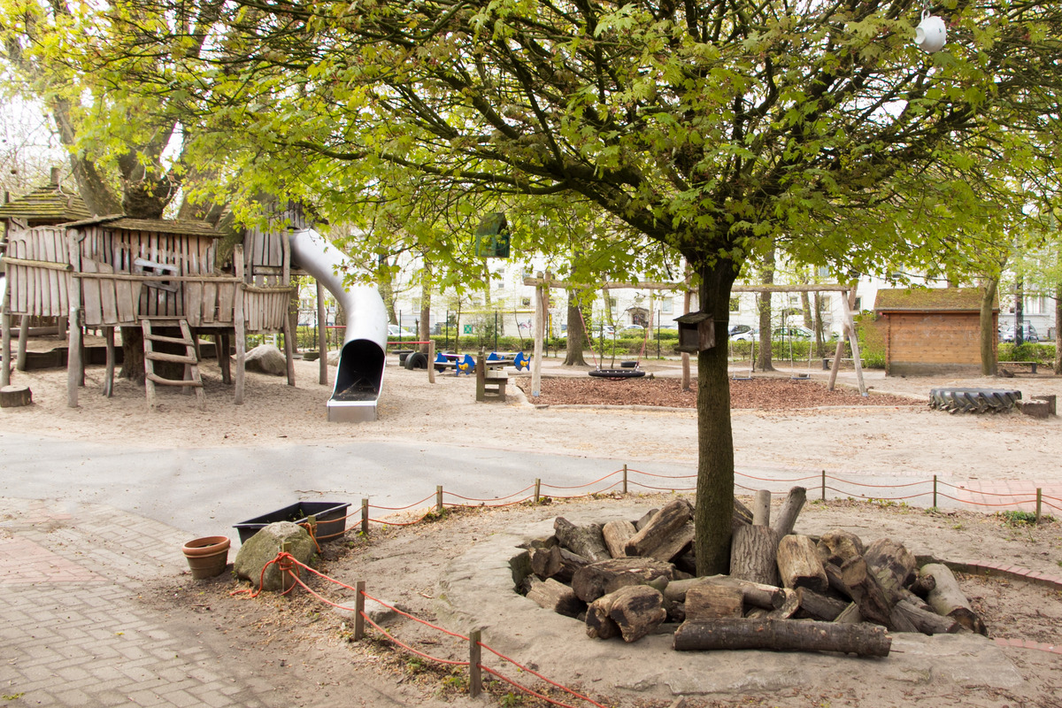 Ein Spielplatz mit einer Holzspielburg, einer Rutsche und einem Baum in der Mitte. Um den Baum liegen Holzstämme. Im Hintergrund sind Schaukeln und eine sandige Fläche. Die Umgebung ist grün und einladend für Kinder.
