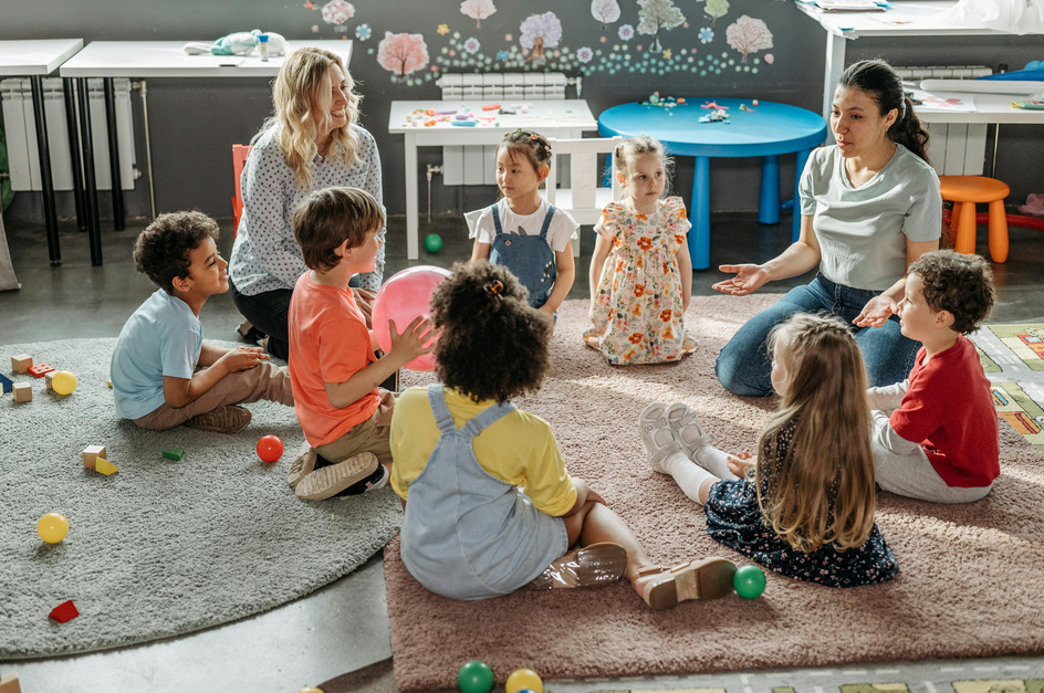 In einem Raum sitzen mehrere Kinder im Kreis auf einem Teppich. Zwei Erzieherinnen sprechen zu ihnen, während einige Kinder aufmerksam zuhören. Spielzeug und bunte Bälle liegen verstreut auf dem Boden. 