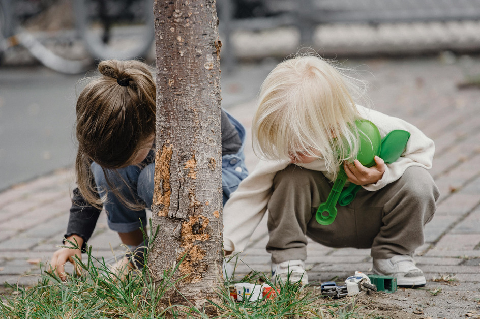 Zwei Kinder hocken neben einem Baum und betrachten den Boden. Um sie herum ist Gras und ein paar kleine Spielzeugautos sind auf dem Boden verteilt.