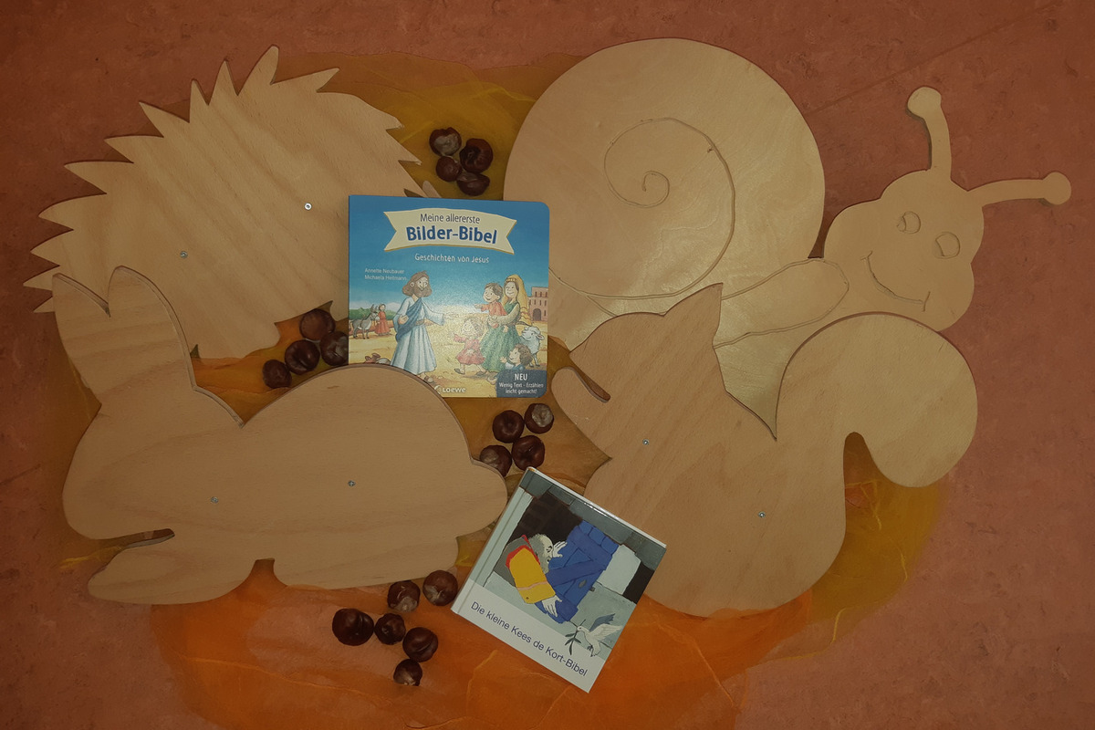 Eine Sammlung von holzgeschnittenen Tieren, darunter ein Igel, ein Hase, ein Eichhörnchen und eine Schnecke, liegt zusammen mit zwei Kinderbüchern. Eines der Bücher ist die „Bilder-Bibel“, das Geschichten von Jesus enthält. 