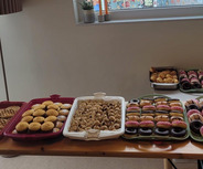 Ein Tisch, der mit verschiedenen Backwaren bedeckt ist, darunter Kekse, Gebäck und bunte Donuts. Im Hintergrund befindet sich ein großes Fenster mit Blick auf eine Straße, auf der ein Geschäftsschild zu sehen ist.