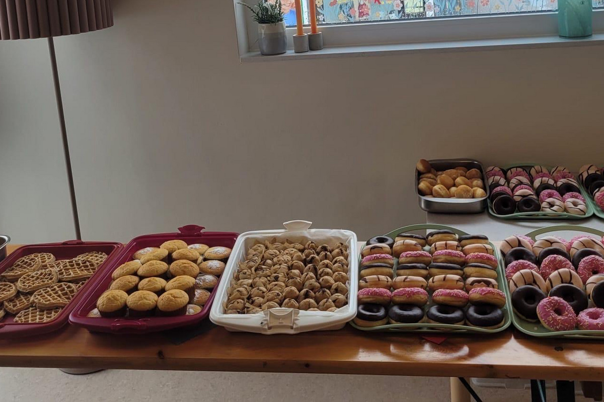Ein Tisch, der mit verschiedenen Backwaren bedeckt ist, darunter Kekse, Gebäck und bunte Donuts. Im Hintergrund befindet sich ein großes Fenster mit Blick auf eine Straße, auf der ein Geschäftsschild zu sehen ist.