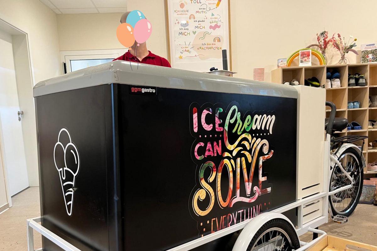 Eine Person steht hinter einem großen Eiswagen mit einem bunten Schild, auf dem „Ice Cream Can Solve Everything“ („Eis kann alles lösen“) steht. Der Eiswagen befindet sich in einem hellen, großzügigen Raum, in dessen Regalen verschiedene Gegenstände ausgestellt sind.