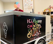 Eine Person steht hinter einem großen Eiswagen mit einem bunten Schild, auf dem „Ice Cream Can Solve Everything“ („Eis kann alles lösen“) steht. Der Eiswagen befindet sich in einem hellen, großzügigen Raum, in dessen Regalen verschiedene Gegenstände ausgestellt sind.