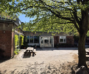 Ein einladender Innenhof mit einem großen, schattenspendenden Baum und einem Tisch. Im Hintergrund steht ein Gebäude mit großen Fenstern und Eingangstüren. Der Himmel ist klar und sonnig, und der Boden ist teilweise mit Sand bedeckt.