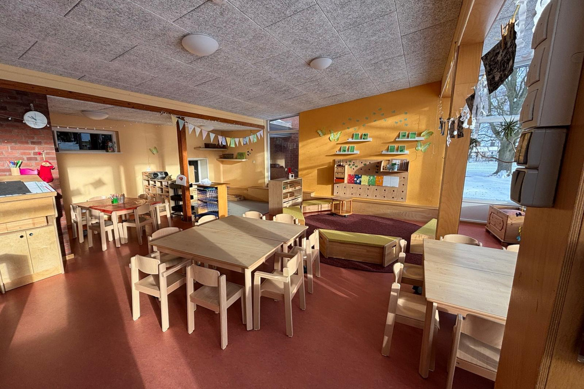 Ein heller, einladender Kindergartenraum mit mehreren Holztischen und -stühlen. An den Wänden sind freundliche Dekorationen und Regale mit Spielmaterialien. Ein gemütlicher Bereich mit einer großen Bank lädt zum Entspannen ein. Licht strömt durch die Fenster, die den Blick auf eine verschneite Landschaft ermöglichen.