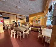 Ein heller, einladender Kindergartenraum mit mehreren Holztischen und -stühlen. An den Wänden sind freundliche Dekorationen und Regale mit Spielmaterialien. Ein gemütlicher Bereich mit einer großen Bank lädt zum Entspannen ein. Licht strömt durch die Fenster, die den Blick auf eine verschneite Landschaft ermöglichen.