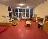 Der Raum ist ein heller Kindergarten mit einem roten Boden. Es gibt verschiedene Möbel: einen Tisch mit Stühlen, Holzregale, Spielzeugkisten und eine kleine Spielbühne. An der Wand hängen bunte Bilder. Große Fenster bieten Blick ins Freie.