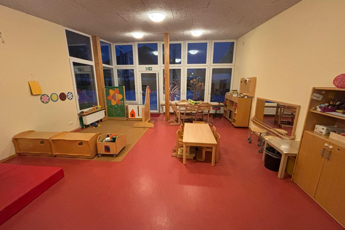 Der Raum ist ein heller Kindergarten mit einem roten Boden. Es gibt verschiedene Möbel: einen Tisch mit Stühlen, Holzregale, Spielzeugkisten und eine kleine Spielbühne. An der Wand hängen bunte Bilder. Große Fenster bieten Blick ins Freie.
