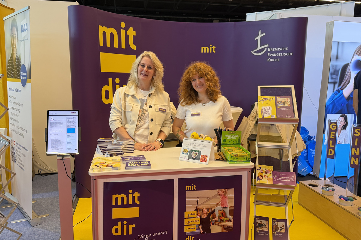Zwei Frauen stehen an einem Messestand mit dem Schriftzug „mit dir“. Der Stand bietet Informationsmaterialien und Broschüren an. Im Hintergrund sind weitere Stände sichtbar.