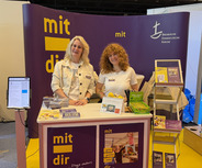 Zwei Frauen stehen an einem Messestand mit dem Schriftzug „mit dir“. Der Stand bietet Informationsmaterialien und Broschüren an. Im Hintergrund sind weitere Stände sichtbar.