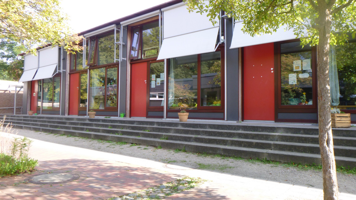 Die Abbildung zeigt die Fassade eines modernen, gebauten Gebäudes mit großen Fenstern in einer roten Wandfarbe. 