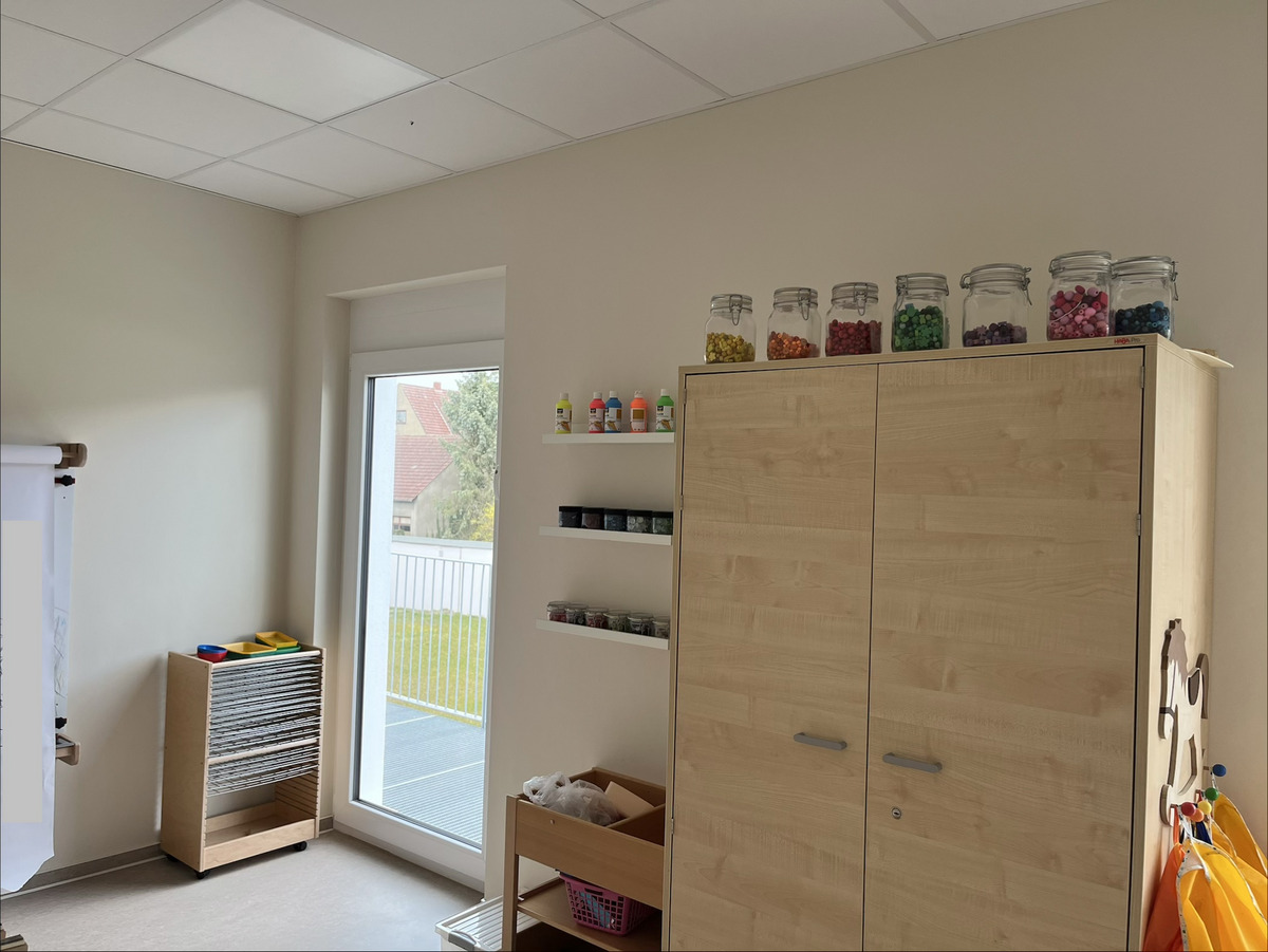 Ein heller Raum mit einem großen Fenster, das nach draußen zeigt. An einer Wand befindet sich ein Holzschrank, auf dem einige Gläser mit bunten Materialien stehen. Neben dem Schrank sind Regale mit Farbflaschen und eine rollbare Holzkommode mit Zeichnungsblättern.