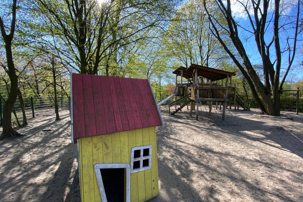 Ein Spielplatz mit einem bunten Spielhaus in Gelb und Rot im Vordergrund. Im Hintergrund steht eine große Holzspielanlage mit einer Rutsche. Umgeben von grünen Bäumen und hellem, sonnigem Himmel. Der Sandboden rundet die spielerische Atmosphäre ab.