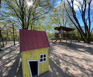 Ein Spielplatz mit einem bunten Spielhaus in Gelb und Rot im Vordergrund. Im Hintergrund steht eine große Holzspielanlage mit einer Rutsche. Umgeben von grünen Bäumen und hellem, sonnigem Himmel. Der Sandboden rundet die spielerische Atmosphäre ab.