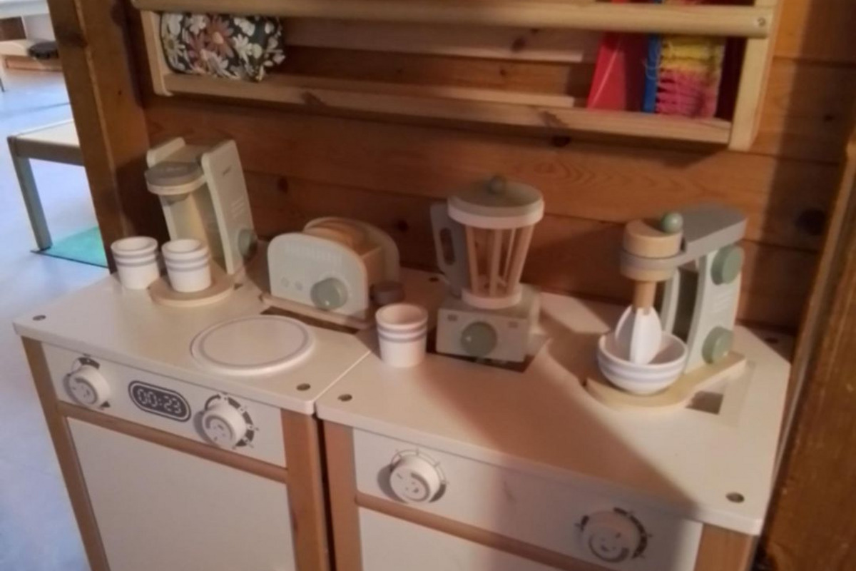 Das Bild zeigt eine Spielküche mit einer Waschmaschine und einem Herd. Auf der Arbeitsfläche stehen Spielzeuggeräte wie ein Mixer, ein Wasserkocher und Tassen. Im Hintergrund sind Reinigungsutensilien wie Schaufel und Besen an einer Stange befestigt. Das gesamte Set ist aus buntem Holz.