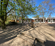 Ein Außenbereich mit einem Sandplatz. Im Vordergrund steht eine Holzbank, daneben ist ein Spielgerät sichtbar. Im Hintergrund sind Bäume und ein Gebäude zu sehen, das große Fenster hat. 