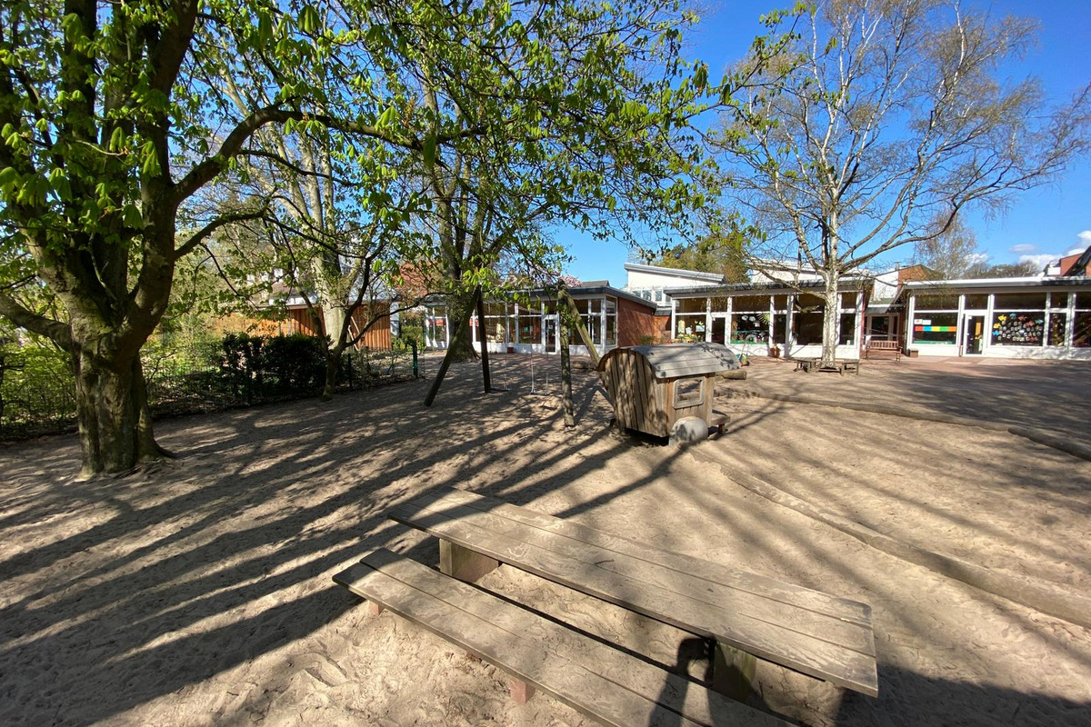 Ein Außenbereich mit einem Sandplatz. Im Vordergrund steht eine Holzbank, daneben ist ein Spielgerät sichtbar. Im Hintergrund sind Bäume und ein Gebäude zu sehen, das große Fenster hat. 