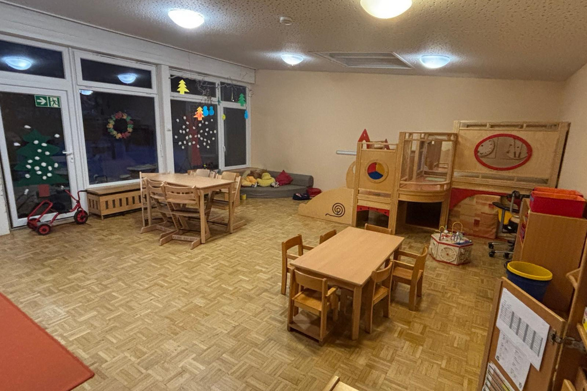 Der Raum zeigt einen Kindergarten mit Holzspielzeug und Tischen. In der Ecke sind bunte Spielgeräte und Kissen. Große Fenster lassen Licht herein, und an den Fenstern hängen Weihnachtsdekorationen. Der Boden ist aus Holzparkett.