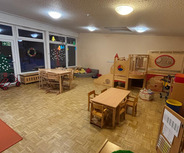 Der Raum zeigt einen Kindergarten mit Holzspielzeug und Tischen. In der Ecke sind bunte Spielgeräte und Kissen. Große Fenster lassen Licht herein, und an den Fenstern hängen Weihnachtsdekorationen. Der Boden ist aus Holzparkett.
