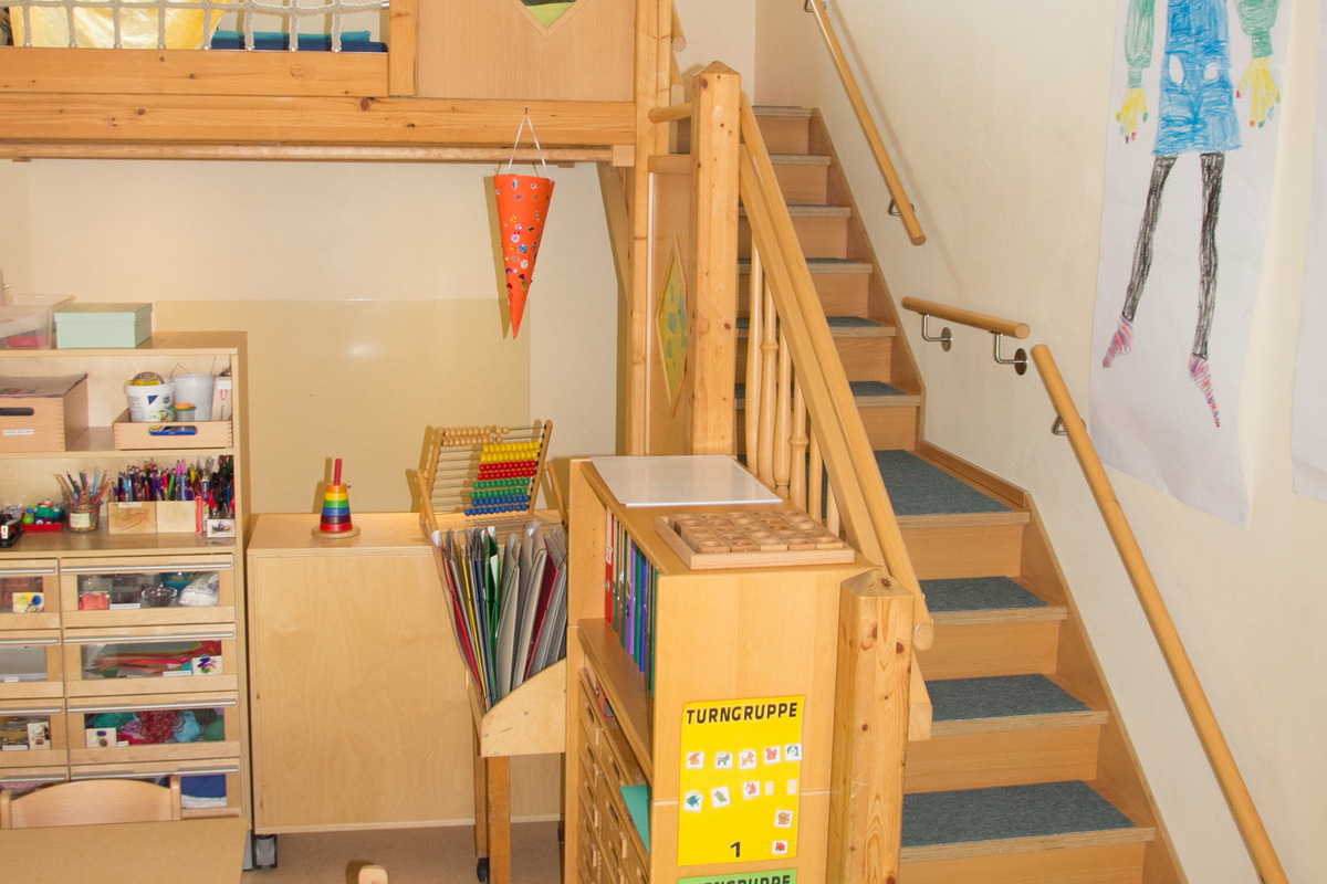 Ein heller Raum mit einem Holzhochbett, von dem eine Treppe nach unten führt. Im Vordergrund stehen Tische und Regale mit Bastelmaterialien. An der Wand hängen bunte Bilder und eine dekorative Papierlaterne. Ein einladender Raum für Kinder zum Spielen und Lernen.