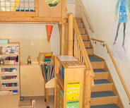 Ein heller Raum mit einem Holzhochbett, von dem eine Treppe nach unten führt. Im Vordergrund stehen Tische und Regale mit Bastelmaterialien. An der Wand hängen bunte Bilder und eine dekorative Papierlaterne. Ein einladender Raum für Kinder zum Spielen und Lernen.