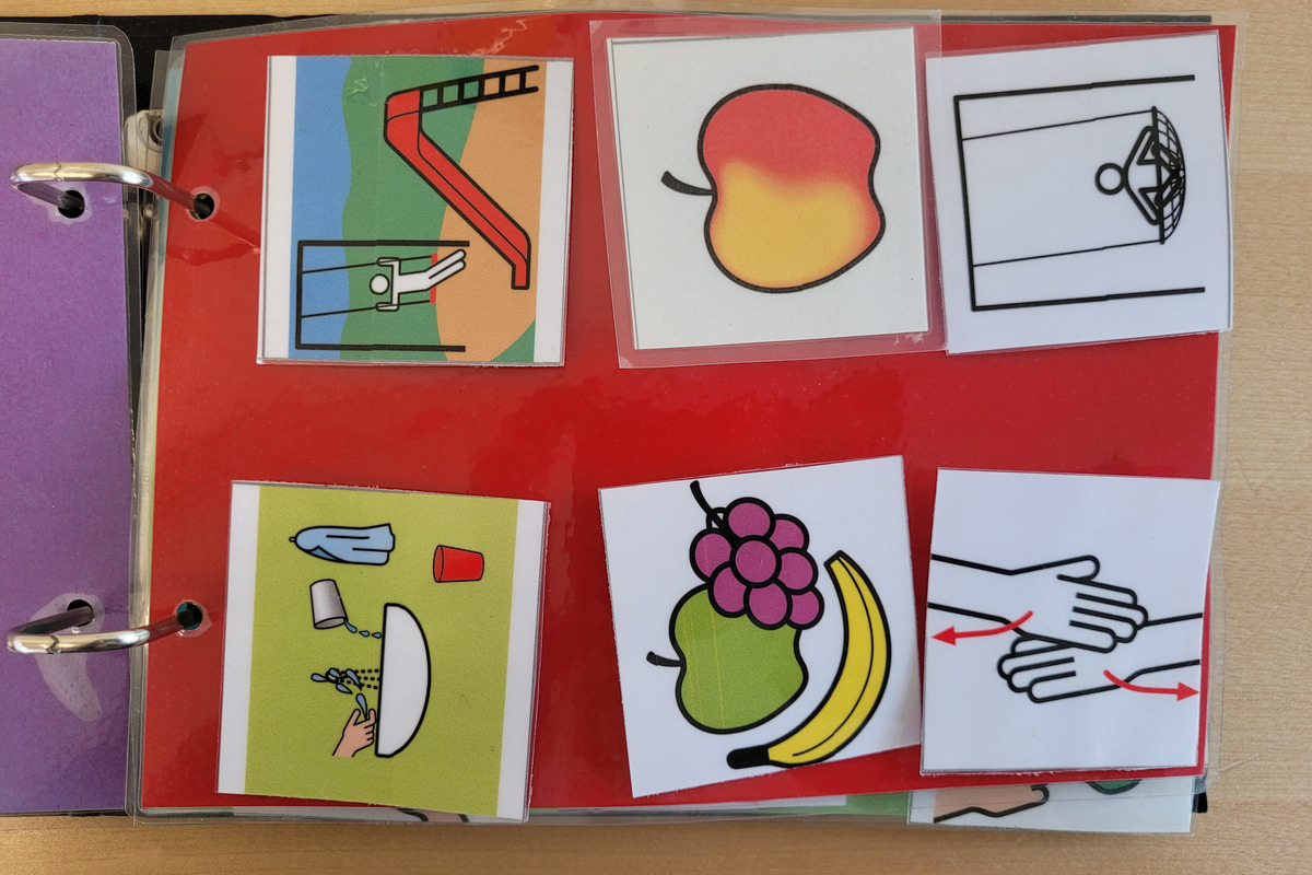 Das Bild zeigt ein buntes Arbeitsheft mit mehreren Symbolen. Darunter sind Bilder von einer Rutsche, einer Frucht, einer Waschaktion und Händen, die sich berühren. Die Symbole können zur Kommunikation oder als Lernhilfen verwendet werden.