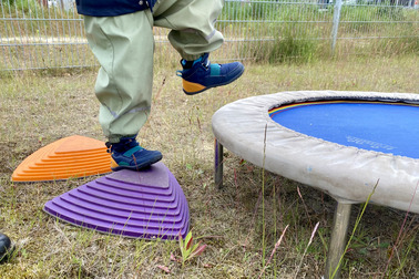 Ein Kind steht auf einem lila Spielgerät und hebt einen Fuß an. Neben dem Spielgerät befindet sich ein Trampolin mit blauer Sprungfläche. Im Hintergrund sind Gras und einige Pflanzen zu sehen.