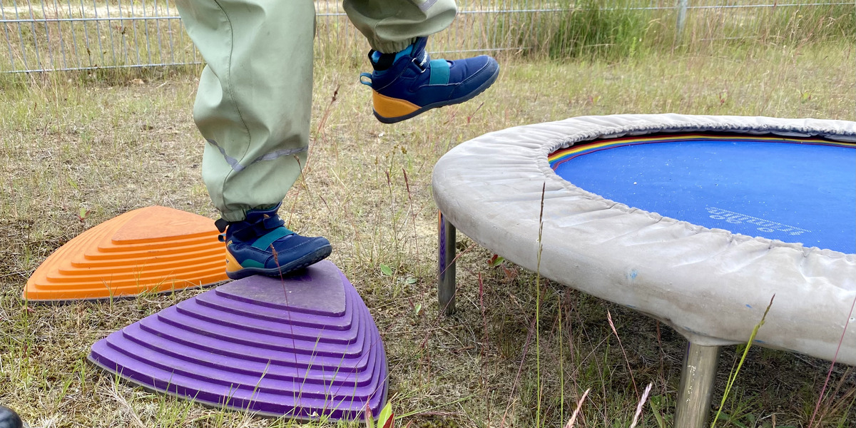 Ein Kind steht auf einem lila Spielgerät und hebt einen Fuß an. Neben dem Spielgerät befindet sich ein Trampolin mit blauer Sprungfläche. Im Hintergrund sind Gras und einige Pflanzen zu sehen.