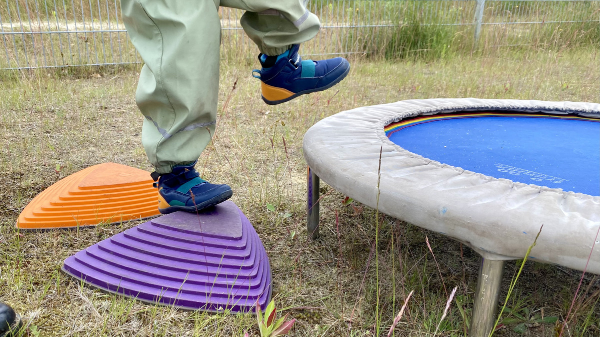 Ein Kind steht auf einem lila Spielgerät und hebt einen Fuß an. Neben dem Spielgerät befindet sich ein Trampolin mit blauer Sprungfläche. Im Hintergrund sind Gras und einige Pflanzen zu sehen.