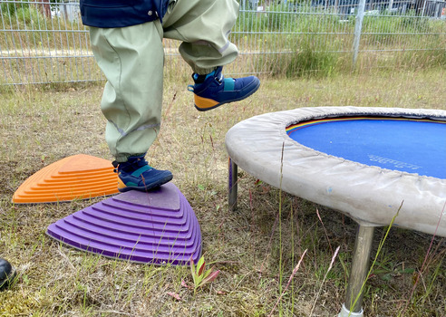 Ein Kind steht auf einem lila Spielgerät und hebt einen Fuß an. Neben dem Spielgerät befindet sich ein Trampolin mit blauer Sprungfläche. Im Hintergrund sind Gras und einige Pflanzen zu sehen.