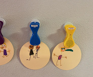 Drei runde, beigen Papierscheiben hängen an der Wand. Jede Scheibe hat eine Zeichnung von einfachen Figuren; die erste zeigt zwei Personen, die zweite zwei weitere in Umarmung, und die dritte zeigt eine stehende Figur mit einer Blume. Über den Scheiben befinden sich Sprachklammern.