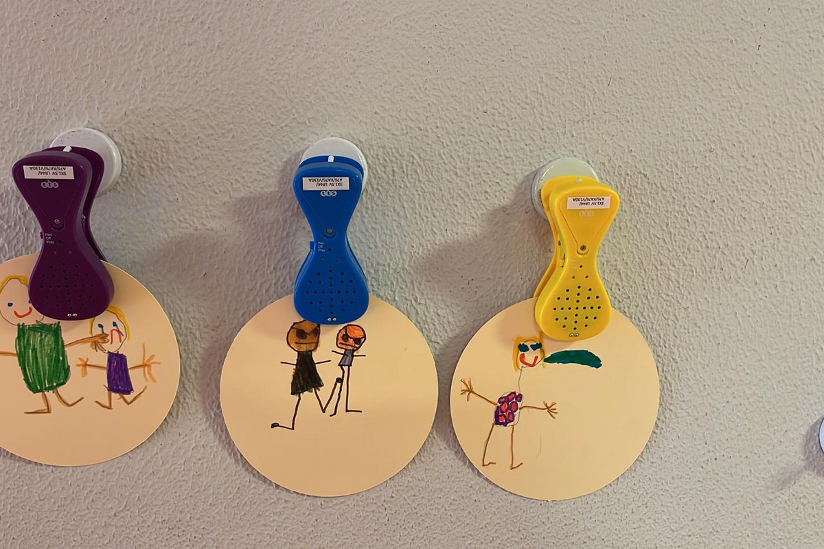 Drei runde, beigen Papierscheiben hängen an der Wand. Jede Scheibe hat eine Zeichnung von einfachen Figuren; die erste zeigt zwei Personen, die zweite zwei weitere in Umarmung, und die dritte zeigt eine stehende Figur mit einer Blume. Über den Scheiben befinden sich Sprachklammern.