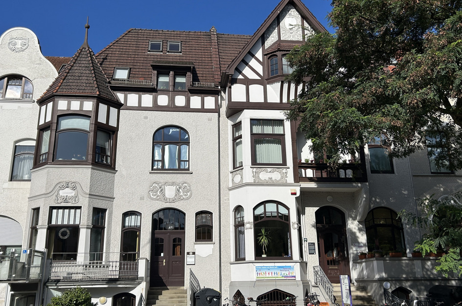 Ein historisches Gebäude mit einem schönen, verzierten Eingangsbereich und großen Fenstern. Die Fassade ist hellgrau mit weißen und braunen Akzenten. Auf der linken Seite ist ein Erkerfenster zu sehen. Vor dem Gebäude stehen Fahrräder und ein kleiner Garten ist sichtbar.