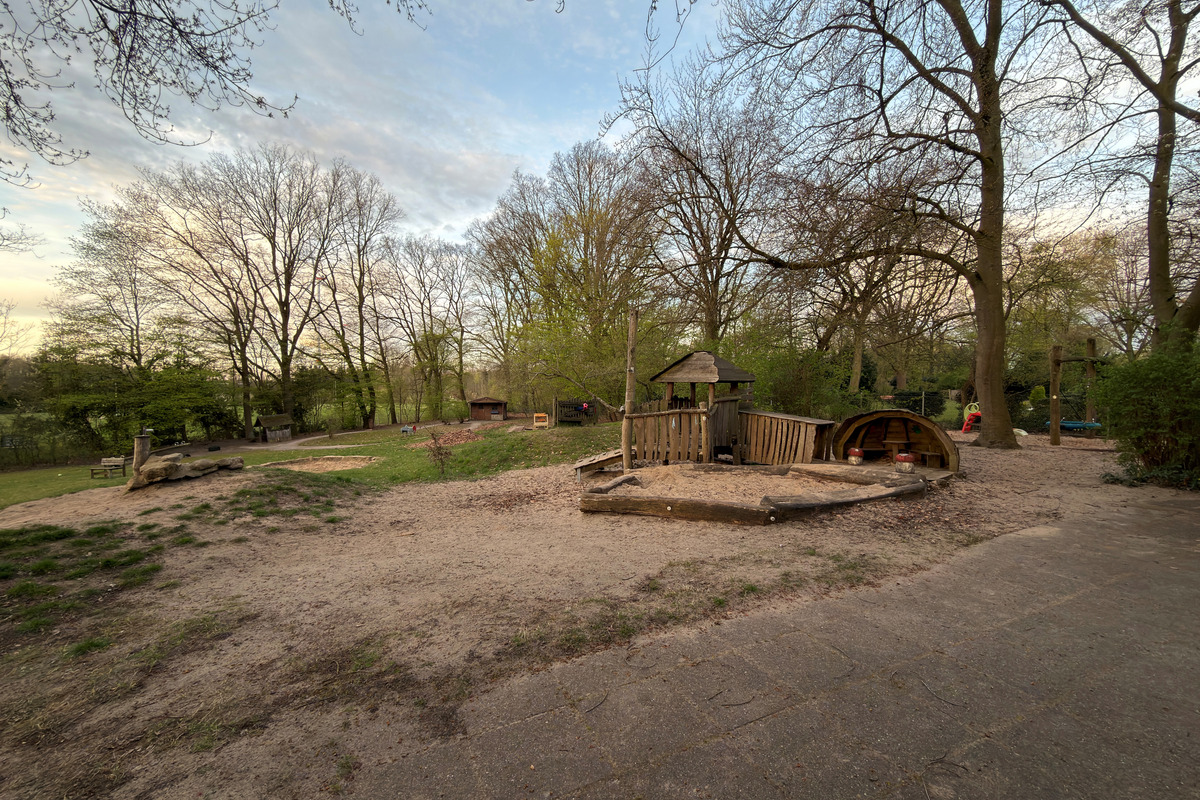 Der Spielplatz zeigt einen Sandbereich mit Spielgeräten aus Holz, darunter ein kleiner Spielturm und eine Bootskonstruktion. Die Umgebung ist von Bäumen umgeben, die frisches Grün tragen. Der Himmel ist teilweise bewölkt, und die Atmosphäre wirkt einladend für Kinder.