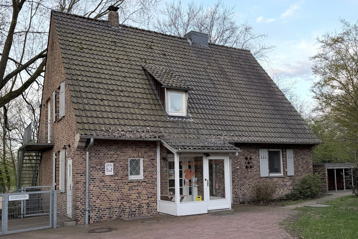 Ein zweigeschössiges, ziegelsteingekleidetes Haus mit einem steilen Dach. Die Vorderseite hat große Fenster und eine gläserne Eingangstür. Vor dem Haus befindet sich ein gepflasterter Weg, umgeben von Bäumen und einer kleinen Grünfläche.