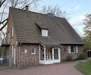 Ein zweigeschössiges, ziegelsteingekleidetes Haus mit einem steilen Dach. Die Vorderseite hat große Fenster und eine gläserne Eingangstür. Vor dem Haus befindet sich ein gepflasterter Weg, umgeben von Bäumen und einer kleinen Grünfläche.