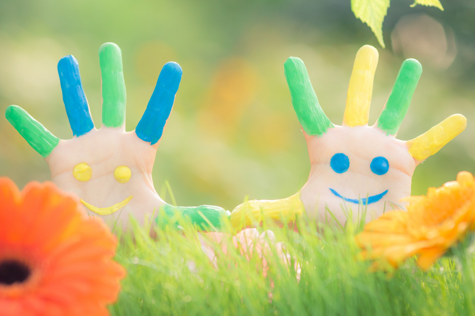 Zwei Hände mit buntem Fingerfarben auf einem grünen Hintergrund. Eine Hand zeigt blau und grün, die andere gelb und orange. Beide haben lächelnde Gesichter mit blauen Augen. Bunte Blumen sind im Vordergrund sichtbar.