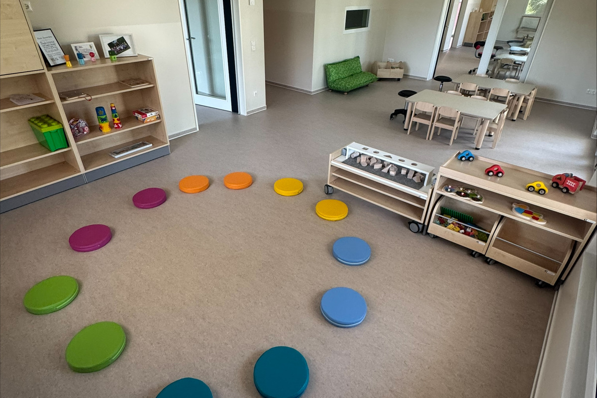 Ein heller Raum mit einer Spielecke. Auf dem Boden liegt ein Kreis aus farbigen Kissen. An einer Seite stehen Holzregale mit Spielzeug und Spieltischen. Im Hintergrund sind Türen und Fenster sichtbar.