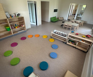 Ein heller Raum mit einer Spielecke. Auf dem Boden liegt ein Kreis aus farbigen Kissen. An einer Seite stehen Holzregale mit Spielzeug und Spieltischen. Im Hintergrund sind Türen und Fenster sichtbar.