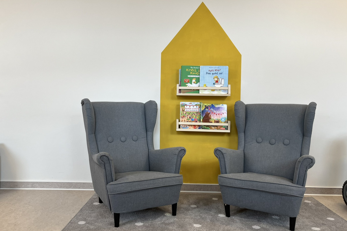 Zwei graue Sessel stehen gegenüber auf einem Teppich mit Punkten. Hinter den Sesseln befindet sich ein Regal mit bunten Kinderbüchern, die an einer gelben Wand hängen.