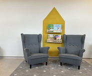 Zwei graue Sessel stehen gegenüber auf einem Teppich mit Punkten. Hinter den Sesseln befindet sich ein Regal mit bunten Kinderbüchern, die an einer gelben Wand hängen.