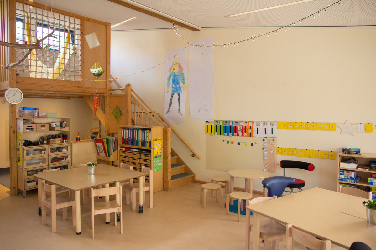 Ein heller, einladender Klassenraum mit Holzmöbeln. Im Raum stehen Tische und Stühle. Eine Treppe führt nach oben. An den Wänden hängen bunte Zeichnungen und Lernmaterialien. Ein Regal ist mit Bastelmaterialien und Büchern gefüllt. Eine kleine Pflanze steht auf einem Tisch.