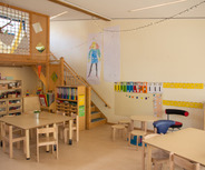 Ein heller, einladender Klassenraum mit Holzmöbeln. Im Raum stehen Tische und Stühle. Eine Treppe führt nach oben. An den Wänden hängen bunte Zeichnungen und Lernmaterialien. Ein Regal ist mit Bastelmaterialien und Büchern gefüllt. Eine kleine Pflanze steht auf einem Tisch.