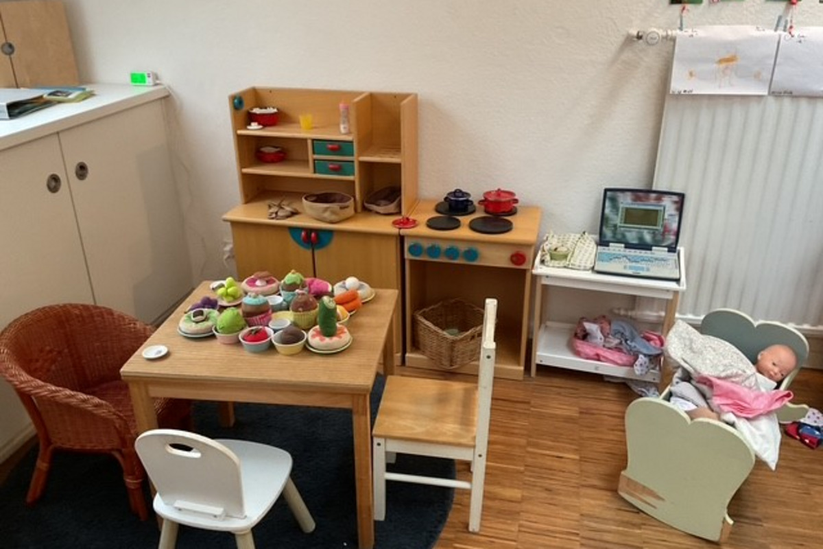 Ein Spielzimmer mit einem kleinen Tisch, Stühlen und einer Spielküche. Der Tisch ist mit Obst- und Gemüsesorten dekoriert. Links steht ein Korb, und rechts ist eine Puppenwiege mit einer Puppe. 