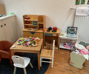 Ein Spielzimmer mit einem kleinen Tisch, Stühlen und einer Spielküche. Der Tisch ist mit Obst- und Gemüsesorten dekoriert. Links steht ein Korb, und rechts ist eine Puppenwiege mit einer Puppe. 