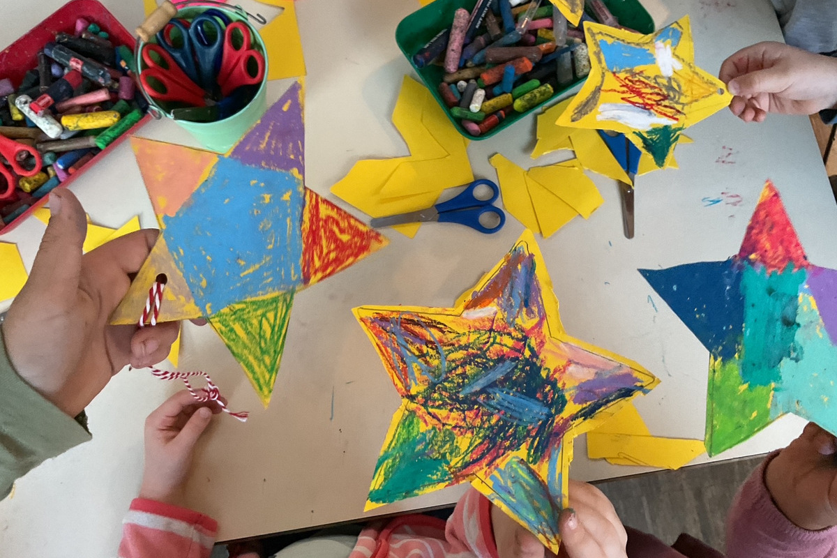 Mehrere Hände halten bunte, gemalte Sterne. Auf einem Tisch liegen weitere Sterne, Scheren, bunten Kreide und Bastelmaterialien. Die Szene zeigt Kinder, die kreativ basteln.