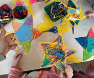 Mehrere Hände halten bunte, gemalte Sterne. Auf einem Tisch liegen weitere Sterne, Scheren, bunten Kreide und Bastelmaterialien. Die Szene zeigt Kinder, die kreativ basteln.