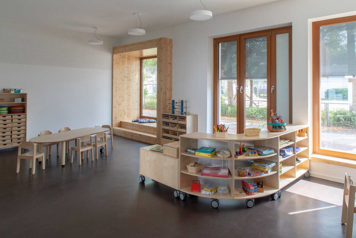 Ein heller, einladender Raum mit einem ovalen Tisch und Stühlen aus Holz. An einer Wand stehen Regale mit Spielzeug und Bastelmaterial.